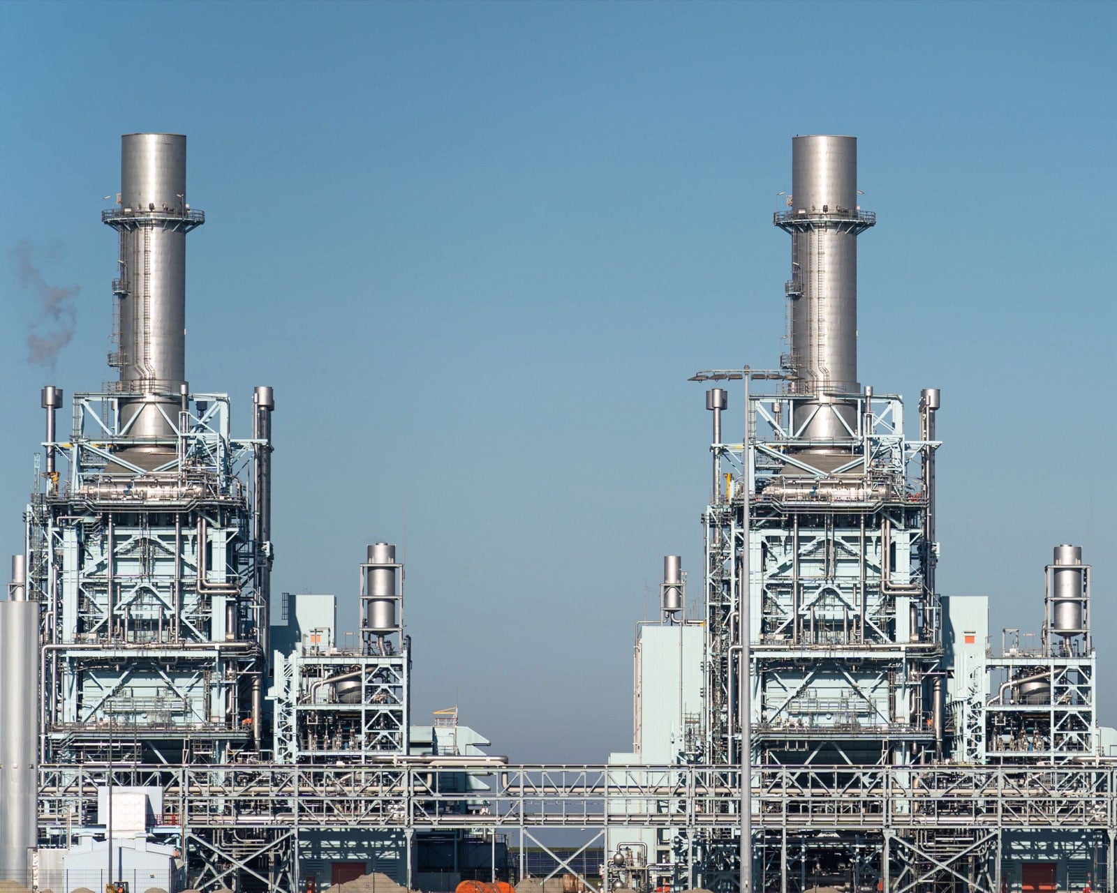 Two gas-fired generating units at a power plant, each with a tall silver exhaust stack above a steel-framed structure, against a clear blue sky